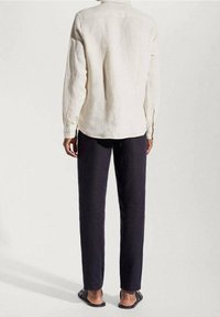 Persona vestita con una camicia bianca a maniche lunghe, pantaloni neri e sandali neri senza lacci, in piedi contro uno sfondo chiaro e semplice.