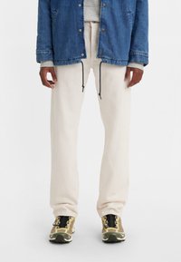 Pantalones beige claros de corte relajado, con cintura ajustable de cordón, combinados con una chaqueta de mezclilla azul y zapatillas deportivas de varios colores.