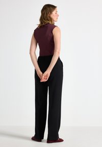 Burgunderfarbener, gerippter ärmelloser Top, kombiniert mit hoch sitzenden schwarzen Hosen, die stilvolle Designdetails aufweisen. Das Model steht mit hinter dem Rücken verschränkten Händen.