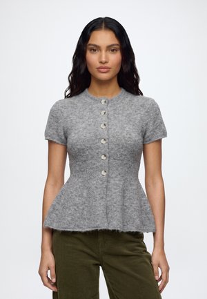Femme aux longs cheveux foncés portant un pull gris à manches courtes boutonné et un pantalon vert olive sur un fond blanc uni.