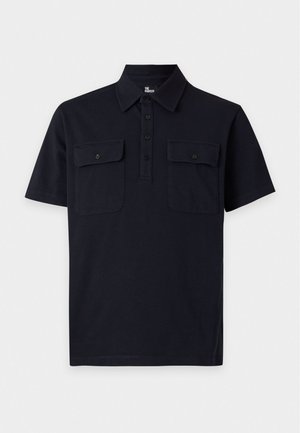 Jeune homme debout portant une chemise noire à manches courtes avec col et deux poches poitrine, un jean bleu clair et des chaussures noires sur un fond uni.