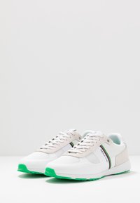 Weiße und hellgraue Sneakers mit grünem Gummisohle, aus Mesh- und Wildledermaterialien, mit einem mehrfarbigen Streifen und Schnürdesign.