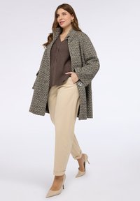 Přesměrovaný šedý kabát s hounds-tooth vzorem vyrobený z texturované látky, velké knoflíky; zkombinovaný s hnědým žebrovaným svetr a béžovými kalhotami s ohrnutými nohavicemi.