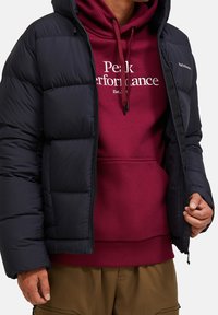Marineblå puffjakke med quiltet design, med frontlynlås og hætte. Underneath en bordeaux sweatshirt med en hvid logo.