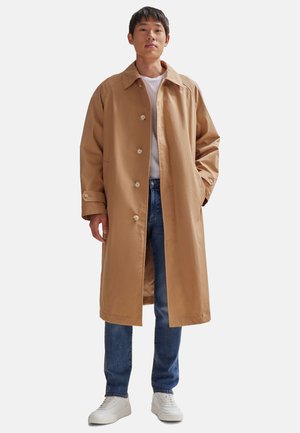 Beige Baumwoll-Trenchcoat mit geradem Schnitt, großem Kragen und Knopfverschluss, kombiniert mit blauen Jeans und weißen Sneakers.