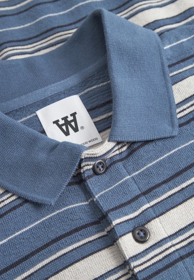 Polo à rayures présentant des couleurs bleu, marine et crème. Tissu en coton au toucher doux, avec une patte de boutonnage et un col. Étiquette de marque visible.