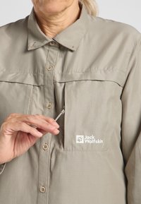 Helle beige Knopfleiste mit Kragen, ausgestattet mit einer Brusttasche und einem Markenlogo. Das Material wirkt glatt und leicht.
