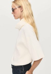 Maglione bianco, oversize, con colletto alto, maniche corte, texture liscia, abbinato a jeans blu scuro e occhiali da sole. Design semplice e moderno.