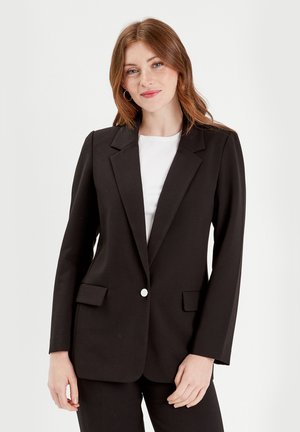 Blazer nero su misura con rever a U, tasche frontali e chiusura con un singolo bottone bianco. Tessuto liscio e silhouette strutturata.