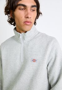 Grå zip-up sweatshirt med høj krave, lavet af blødt bomuldsblandingsstof, med et lille logo-patch på venstre bryst.