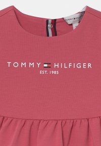 Różowa bawełniana sukienka z marszczonym pasem i okrągłym dekoltem. Z przodu widnieje logo "TOMMY HILFIGER" z rokiem "EST. 1985" poniżej.