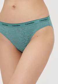 Svetlo teal bikini-style spodnje perilo Calvin Klein je zasnovano s polprosojnim čipkastim vzorcem in črnimi logotipnimi trakovi okoli pasu.