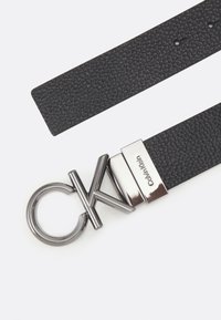 Must matte mustriga vöö, millel on hõbedane CK logo kinnitusega, millel on ringikujuline disain ja metallaktsendid, täiustatud brändingu detailidega.