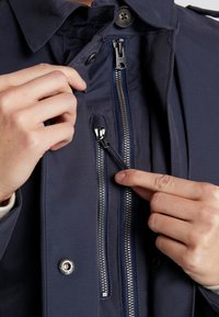 Mains ajustant deux fermetures éclair métalliques verticales sur une veste bleu marine avec un col boutonné.