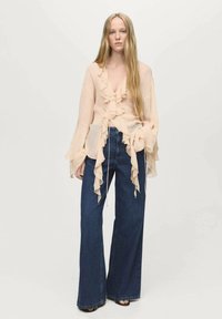 Blouse en pêches à volants avec des manches longues, associée à un jean en denim bleu à taille haute et jambe large. Texture de tissu lisse et design drapé.