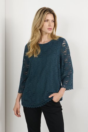 Teal lace top with three-quarter sleeves, scalloped hem, and floral pattern. Worn over black pants with a relaxed fit.
