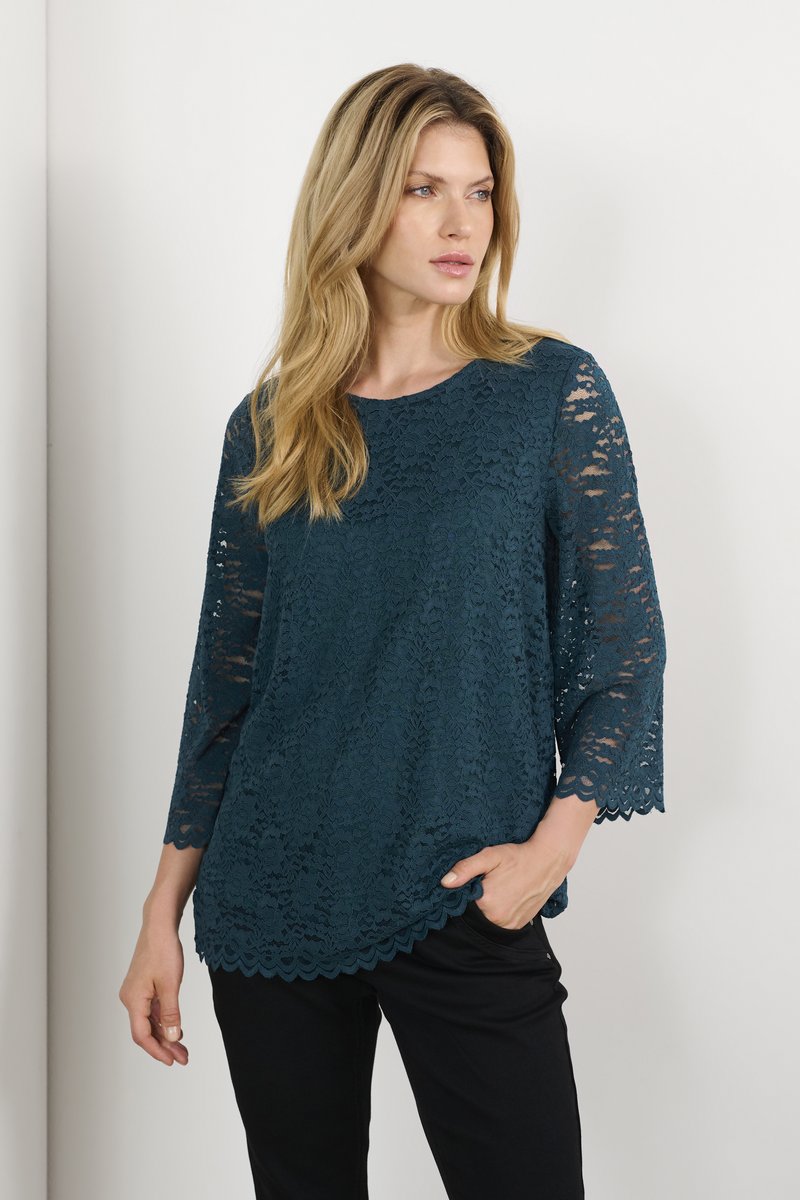 Top in pizzo teal con maniche a tre quarti, orlo scallop e motivo floreale. Indossato sopra pantaloni neri con vestibilità comoda.
