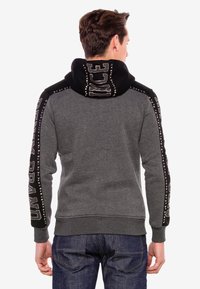 Grauer Hoodie mit schwarzem Kapuzen, versehen mit bedruckten Designs auf den Ärmeln und strukturierten Akzenten an den Kanten. Aus einem weichen Stoff gefertigt.
