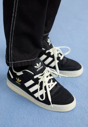 Personne portant des baskets noires Adidas avec des bandes blanches et des lacets, debout sur un sol recouvert d'un tapis bleu clair.