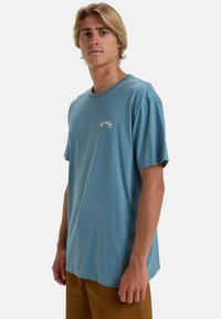 Lys blå bomulds t-shirt med rund hals, korte ærmer og et lille hvidt logo på venstre brystområde. Glat tekstur.