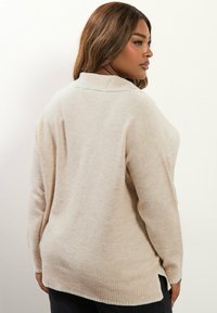 Beige neulepaita, jolla on rento istuvuus, ribbireunat ja sivuhalkiot. Paita sisältää pienen kauluksen ja pehmeän tekstuurin.