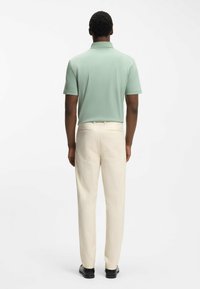 Mand vendt v�æk iført en lysegrøn kortærmet poloshirt, cremefarvede bukser og sorte pæne sko på en ensartet hvid baggrund.