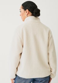 Creme fleece pullover med høj krave og lange ærmer, der har en blød, tekstureret overflade og løst fit, parret med blå jeans.
