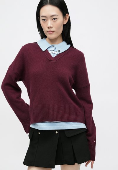 Burgundi kötött pulóver V-nyakkal, világoskék, csíkos galléros inggel párosítva. Fekete redős szoknya zsebekkel.