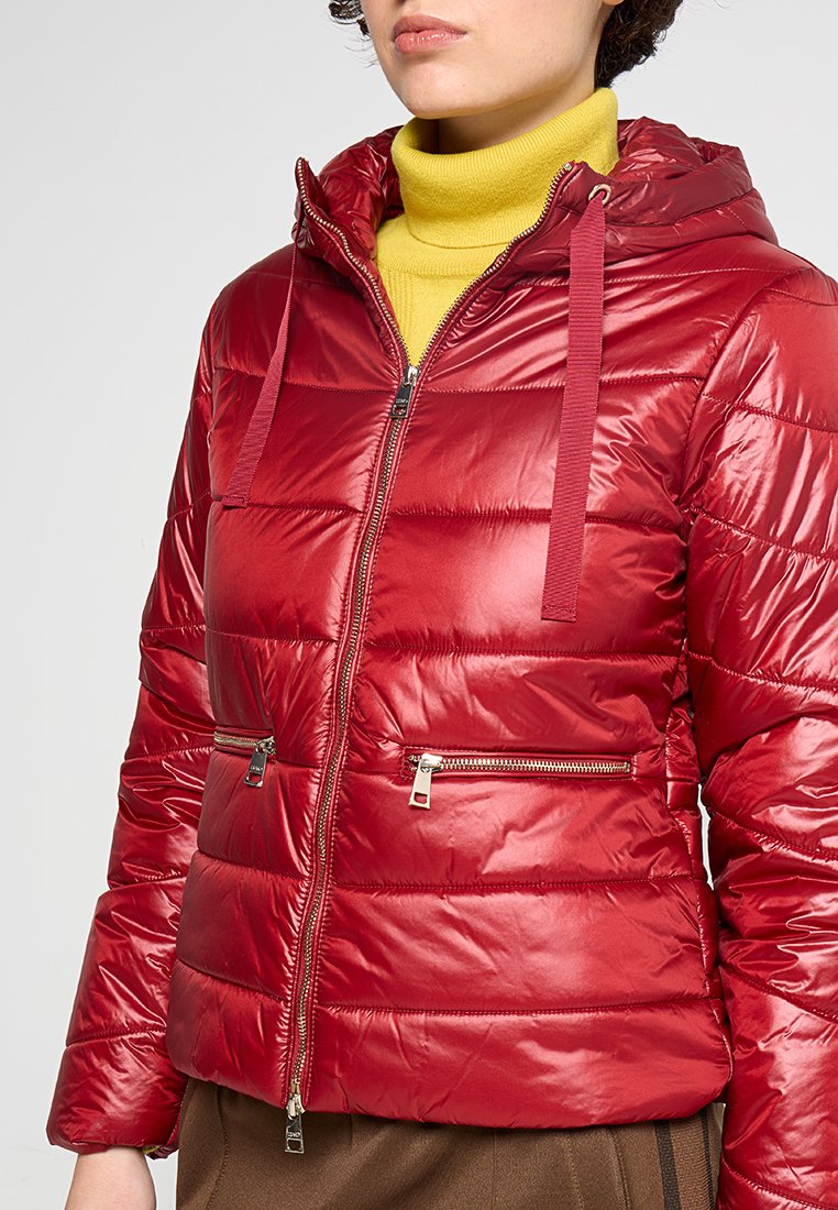 Rood gewatteerd jack met een glanzende afwerking, capuchon, ritssluiting aan de voorkant en twee met rits afgesloten zakken. Het interieur heeft een felgeel coltrui.