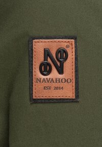 Tejido verde oscuro que presenta un parche de cuero cuadrado con el logotipo en relieve "NAVAHOO" y "EST 2014" en letras negras.