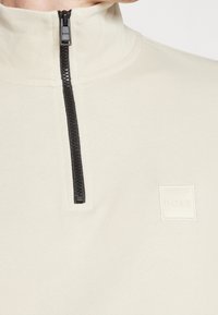 Světle krémový pulover se čtvrtinovým zipem vyrobený z měkké tkaniny, s černým zipem a decentním embosovaným čtvercovým logem na hrudi.