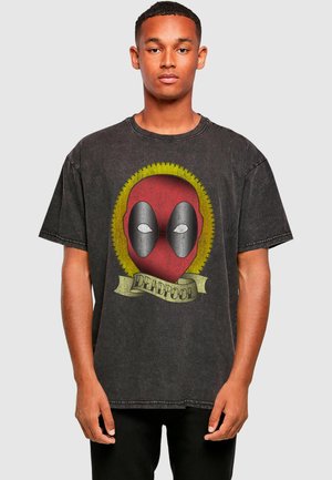 Fekete grafikás póló pamutból, stilizált Deadpool arccal, fehér szemekkel, sárga kör alakú mintával és feliratos szalaggal körülvéve.