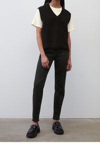 Schwarzes gestricktes ärmelloses Oberteil über einem cremefarbenen T-Shirt, kombiniert mit taillierten schwarzen Hosen und klobigen schwarzen Slippern. Einfärbige Farben, minimalistisches Design.