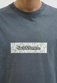 Grå crewneck t-shirt med et tekstureret etikette, der præsenterer logoet "Jack&Jones" på et mønstret baggrund af blade i hvid og gul.