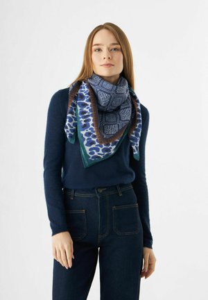 Quadratisches Tuch mit blauen geometrischen und Tiermustern, umrandet von einem braunen Rand, um den Hals drapiert und ergänzt durch einen marineblauen Pullover.
