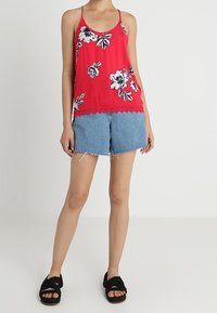 Top corto de flores rojas con tirantes finos, combinado con pantalones cortos de mezclilla azul claro con dobladillos deshilachados. Sandalias negras completan el atuendo.