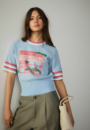Young woman wearing light blue knit top with flower shop illustration, olive pants, and holding a woven beige handbag against a plain wall.
