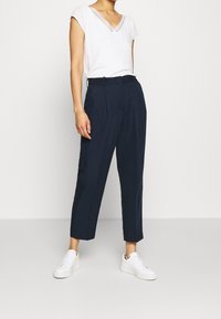 Valkoinen lyhythihainen paita V-päällä ja pitsikoristeluilla, yhdistettynä tummansinisiin leveälahkeisiin housuihin ja valkoisiin tennareihin.