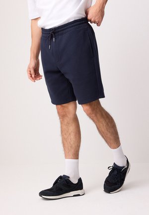 Hombre con pantalones cortos azul marino con cordón, calcetines blancos hasta el tobillo, zapatillas negras y una camisa blanca, de pie contra un fondo claro y sencillo.