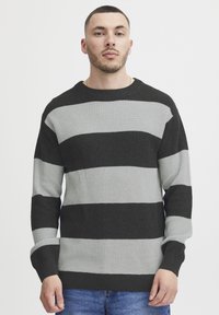 Pulover tricotat cu dungi orizontale îndrăznețe negre și gri, textură ribbed, mâneci lungi și guler rotund.