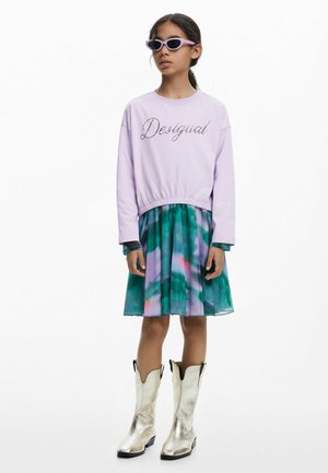 Sudadera color lila con estampado de "Desigual", combinada con un vestido de tie-dye en verde y púrpura. Lleva botas de cowboy plateadas brillantes y gafas de sol.