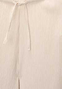 Beżowe spodnie z marszczonego materiału z elastycznym paskiem i subtelnym szwem rozszczepionym w pobliżu dołu.