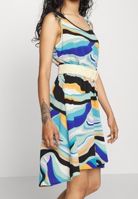A sleeveless dress featuring a multicolored wave pattern in blue, black, yellow, and white, cinched at the waist with a woven beige belt.