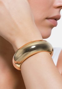 Gold- und roségoldfarbener, metallischer Armreif mit einer glatten, polierten Oberfläche; breite, abgerundete Form, die eng am Handgelenk sitzt.