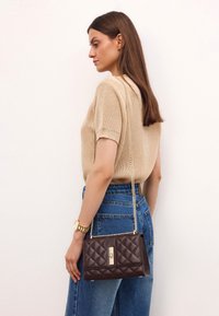 Borsa a tracolla marrone trapuntata con catena dorata e chiusura, in pelle con texture, forma rettangolare. Indossata con un top beige e jeans a vita alta.