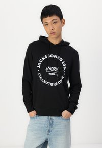 Schwarzer Hoodie aus Baumwolle mit einem Frontdruck, der "JACK&JONES 1990 COLLECTORS CREW" in weißem Text zeigt. Eingestimmt mit hellblauen Jeans.