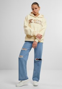 Felpa giallo chiaro con testo grafico marrone a contrasto, abbinata a jeans blu strappati e sneakers bianche. Design semplice e rilassato.
