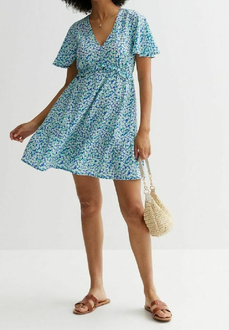 Modré květinové šaty s krátkými rukávy, výstřihem do V, nařaseným pasem a rozšířenou sukňou. Nošeny s hnědými sandály a s pletenou kabelkou na rameno.