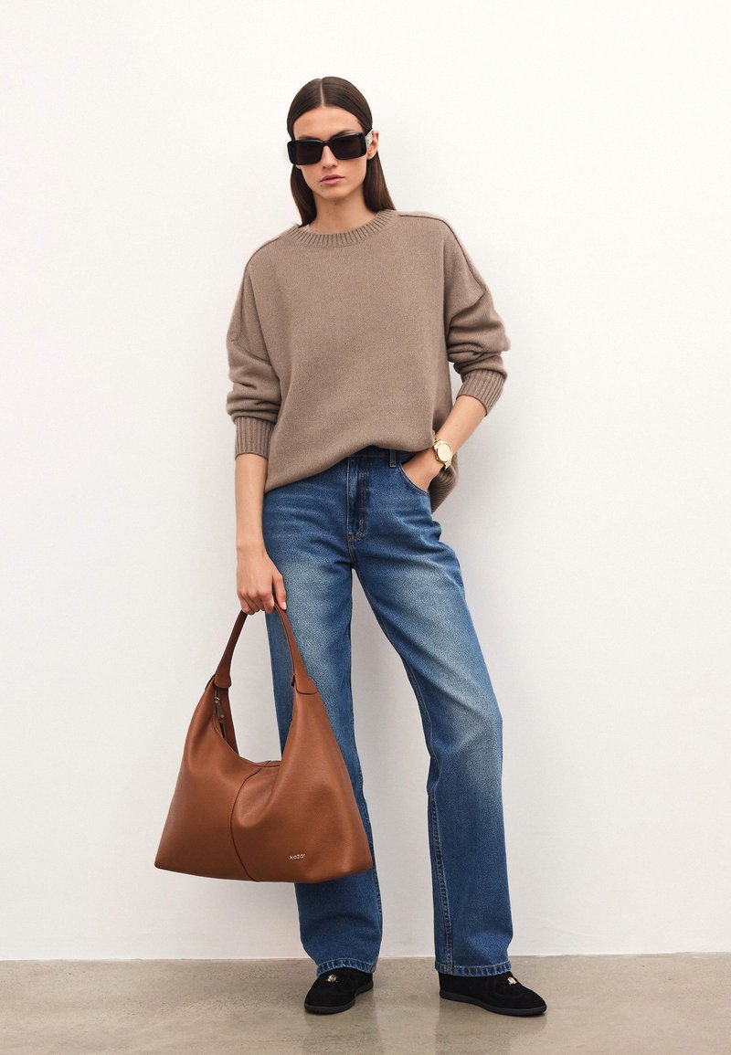 Sieviete ar gariem brūniem matiem, kas valkā lielu bēša džemperi, zila džinsus, melnas kurpes, lielu brūnu somu, melnās saulesbrilles un zelta rokas pulksteni.