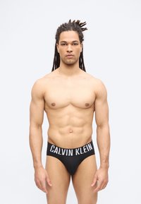 Άνδρας με dreadlocks που φοράει μαύρα εσώρουχα Calvin Klein, στέκεται με το πρόσωπο προς τα εμπρός μπροστά σε ένα απλό φωτεινό φόντο.